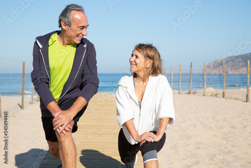 Close-up of sporty elderly couple training with pleasure. Happy man and woman doing sports near blue sea stretching legs smiling looking at each other. Leisure and active life of aged people concept