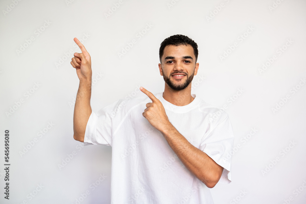 Portrait of positive young man pointing forefingers upwards. Bearded man wearing white T-shirt looking at camera and smiling over white background. Advertising concept Portrait of positive young man pointing forefingers upwards. Bearded man wearing white T-shirt looking at camera and smiling over white background. Advertising concept