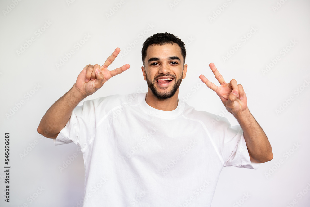 Portrait of playful young man with two victory signs. Happy Caucasian male model with dark hair and beard in white T-shirt looking at camera with tongue out and smiling. Happiness concept Portrait of playful young man with two victory signs. Happy Caucasian male model with dark hair and beard in white T-shirt looking at camera with tongue out and smiling. Happiness concept