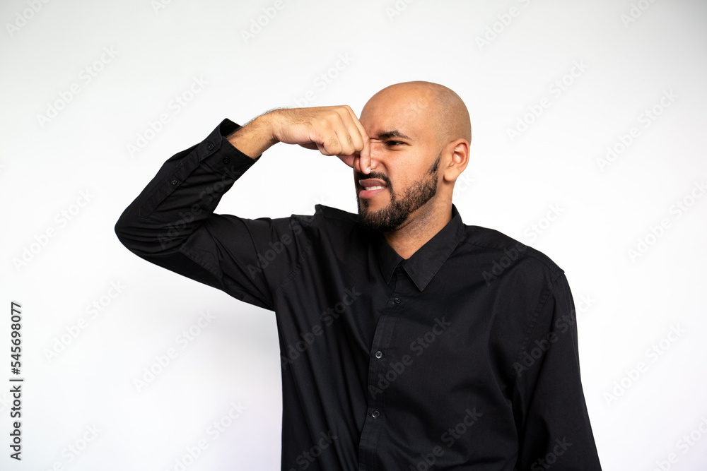 Portrait of young man pinching nose of bad odor over white background. Bearded guy wearing black shirt smelling something with disgust. Stink concept