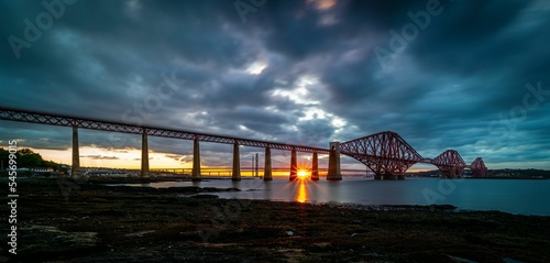 Bright sun shining through Forth Bridge under dark blue sunset clouds in Edinburgh photo