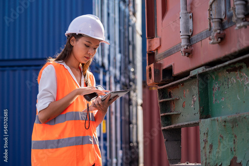 Smile Asian woman logistics worker wear safety helmet and protect suite working and checking product with taplets and scan code gun in shipping containerin commercial transport port