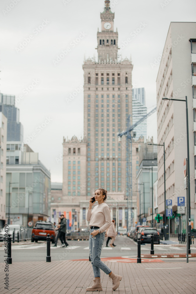 Young woman using mobile phone walking on the city street in Warsaw, Poland. Successful woman calling with cell telephone outside,business talking via mobile phone and walking on the city street.