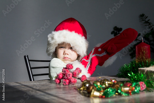 A small child in a Santa Claus hat and red mittens with Christmas gifts is sitting at the New Year's table. Celebrating Christmas.
