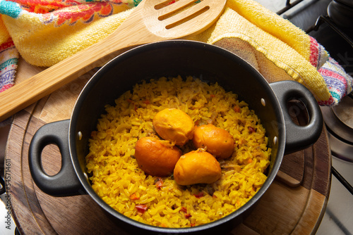 Uma panela preta com arroz com pequi, um pano de prato e uma colher de madeira, sobre uma tábua de madeira. Um prato típico brasileiro.