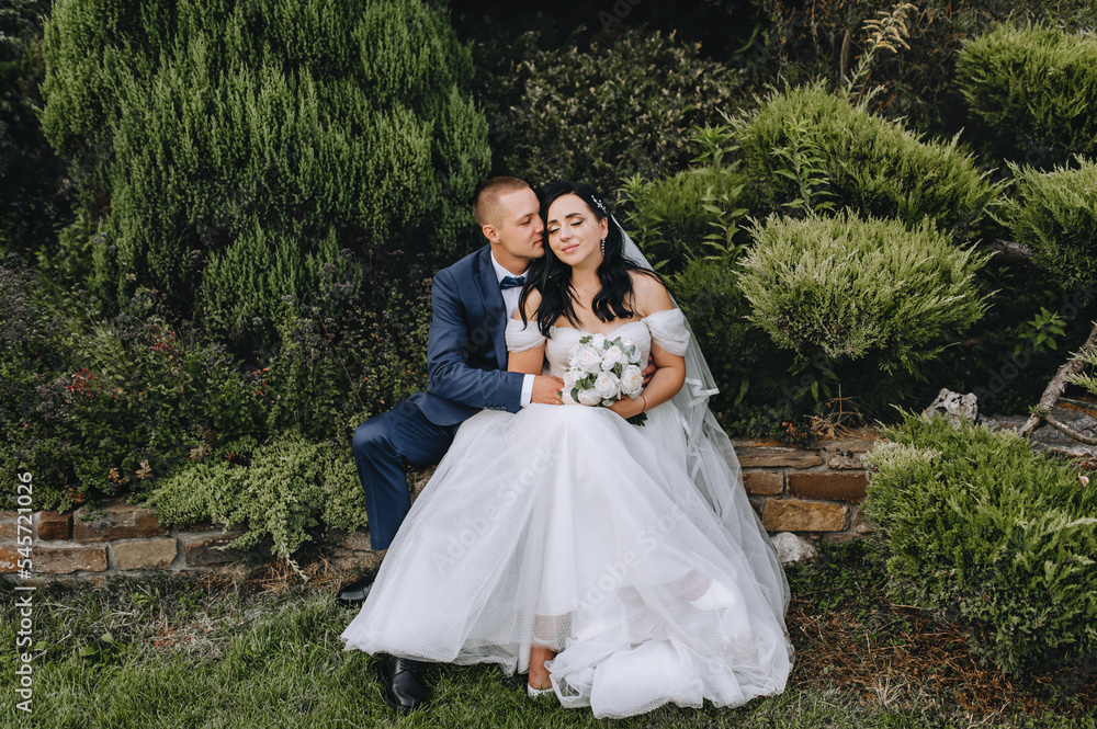 A stylish, young groom in a blue suit and a beautiful, curly brunette bride in a white lace dress with a bouquet gently embrace while sitting in a park outdoors. Wedding photography, portrait.
