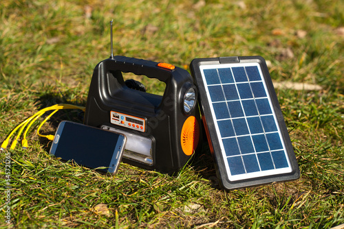 The solar panel charges the battery to which the phone is connected. A telephone, a power bank and a solar panel stand on green grass. Power station for hiking in the mountains