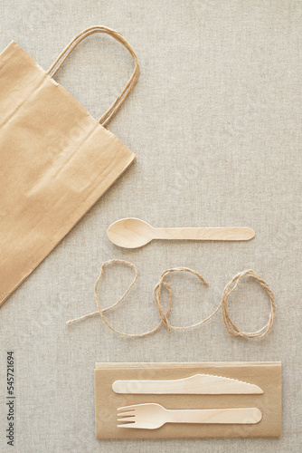 Topic of eco friendly, disposable, recycling, takeaway, plastic free. Craft paper bag, wooden spoon, knife, fork, paper napkin, word eco written with twine on canvas tablecloth. Top view.. Vertical
