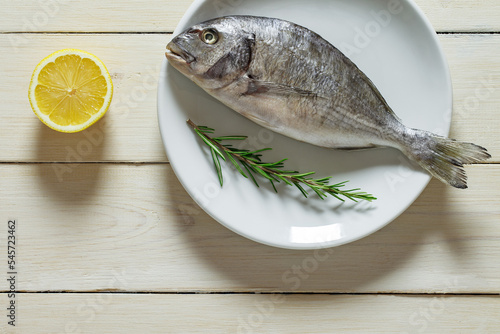 Dorado, raw fresh uncooked fish, cut lemon, rosemary, on plate, on white wooden board background, top view, space to copy text.