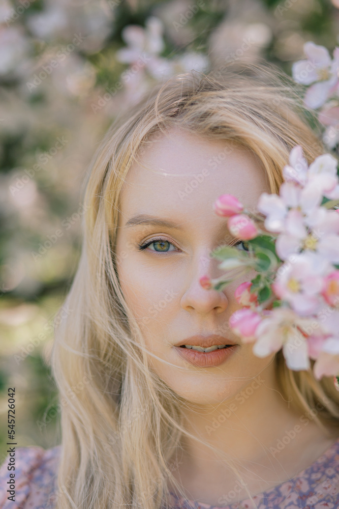Fototapeta premium Portrait of a beautiful girl looking directly at the camera, blooming apple orchard, long blond hair, spring time