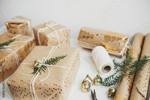 Merry Christmas and Happy Holidays! Stylish christmas gifts. Modern christmas gift boxes with fir branch, golden bell, festive paper and ornaments composition on white wood. Atmospheric time