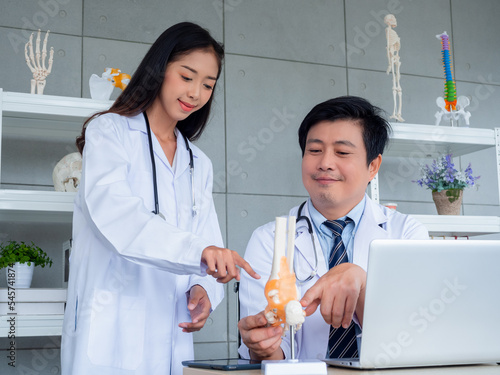 Two Asian doctors in white uniforms talking and working together in medical office. The adult male doctor counseling to young female doctor about the ligaments around the ankle  bone model on desk.