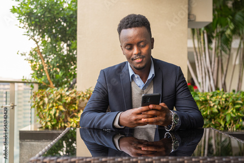 Portrait of handsome black African businessman sitting and using mobile phone