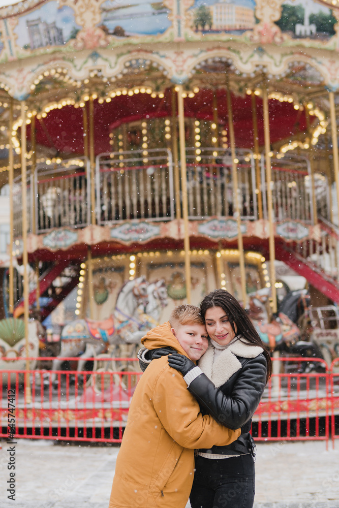 Obraz premium Caucasian mother and son in warm cozy clothes walk outdoors on festive city streets. Family walks at the fair as snow falls. Cold happy winter day. Holidays, Christmas, New Year, love concept.