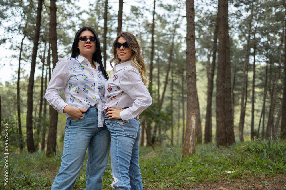 Two model girls who wears same clothes are in forest . White shirt, jean and white boots. One of the is blonde, one of them has black hair. Model shoot. Fashion concept. Two model girls who wears same clothes are in forest . White shirt, jean and white boots. One of the is blonde, one of them has black hair. Model shoot. Fashion concept.
