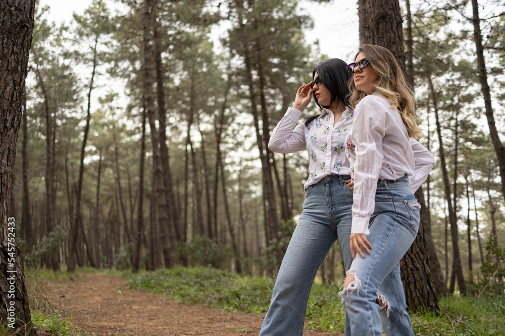 Two model girls who wears same clothes are in forest . White shirt, jean and white boots. One of the is blonde, one of them has black hair. Model shoot. Fashion concept.