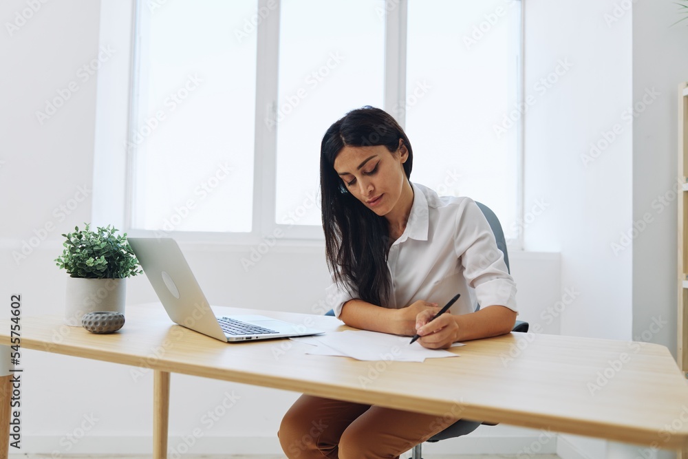 A woman works and learns by writing down text on paper with a pen and checking for errors, studying and teaching in college, business work at a desk with a laptop A woman works and learns by writing down text on paper with a pen and checking for errors, studying and teaching in college, business work at a desk with a laptop