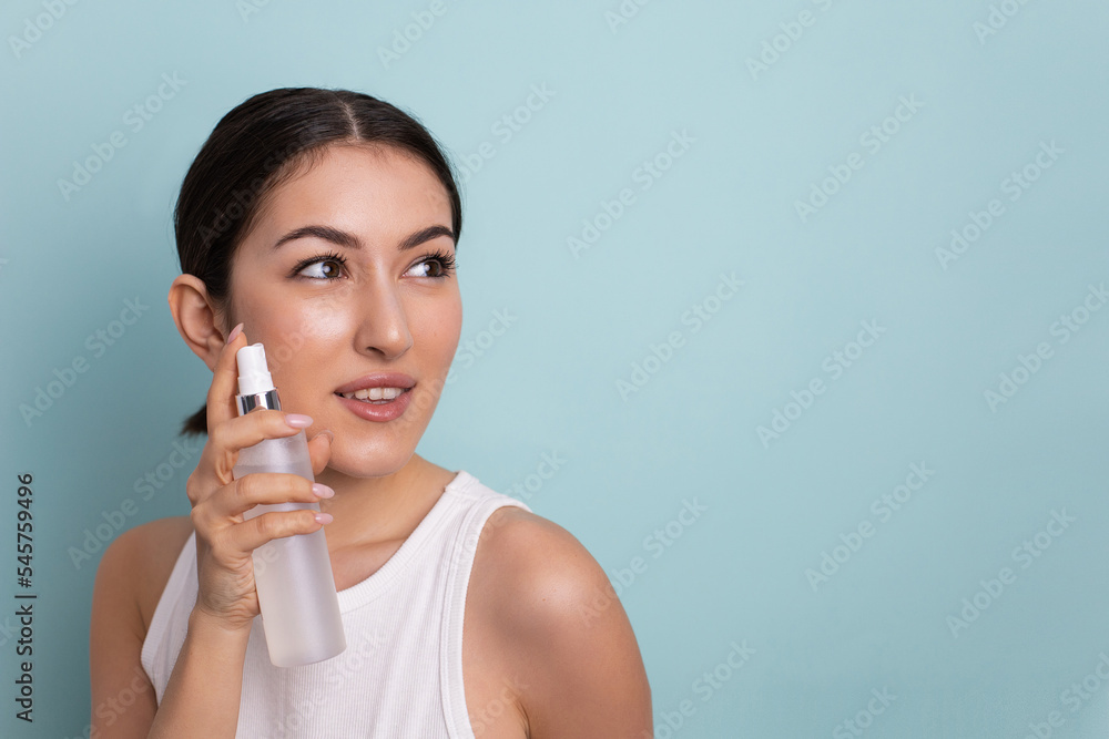 portrait of a beautiful young woman holding a bottle in her hand and spraying liquid with hydration on her face for rejuvenation on a blue background in the studio. portrait of a beautiful young woman holding a bottle in her hand and spraying liquid with hydration on her face for rejuvenation on a blue background in the studio.