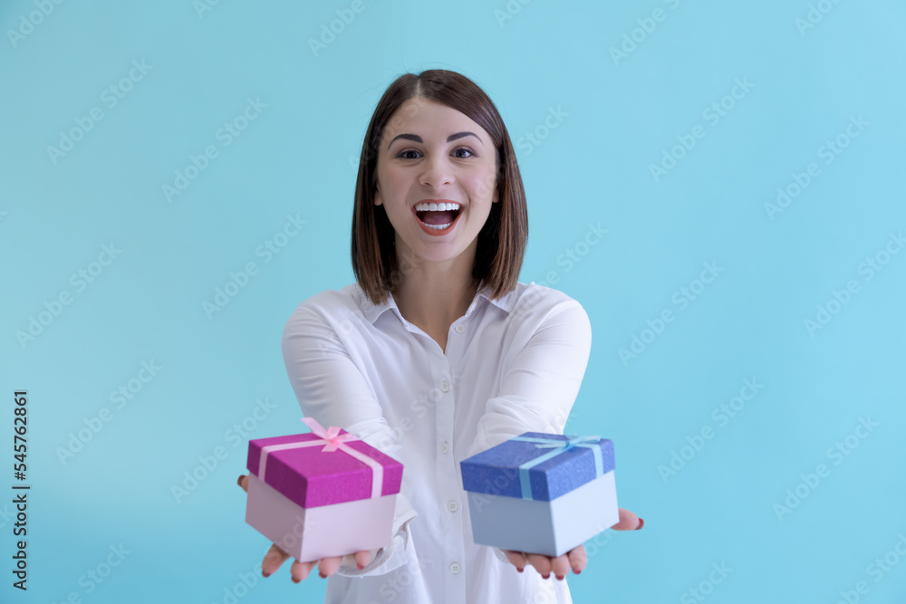 Obraz premium Portrait of happy beautiful brunette woman in white shirt holding pink and blue gift boxes against blue background. Selective focus. Holiday present theme.