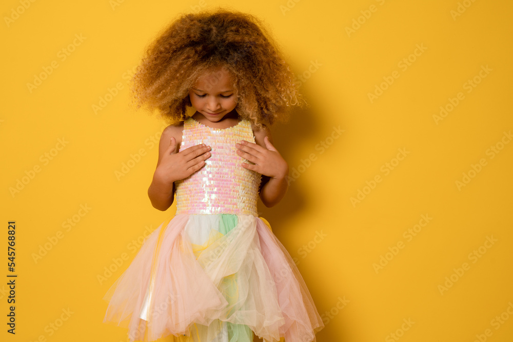 Child girl looking at her cute festive dress standing isolated over yellow background.