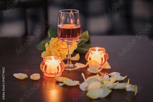 Φωτογραφία A glass of rose wine, two candles and rose petals on a dark table