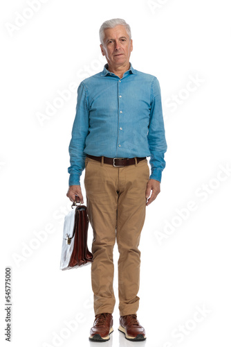 serious old guy with bag wearing denim shirt and chino pants and standing