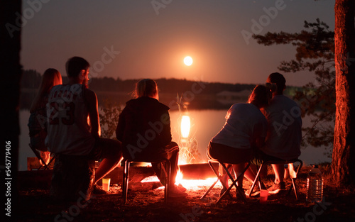 A company of people sits near a fire at night against the backdrop of a lake and the moon.