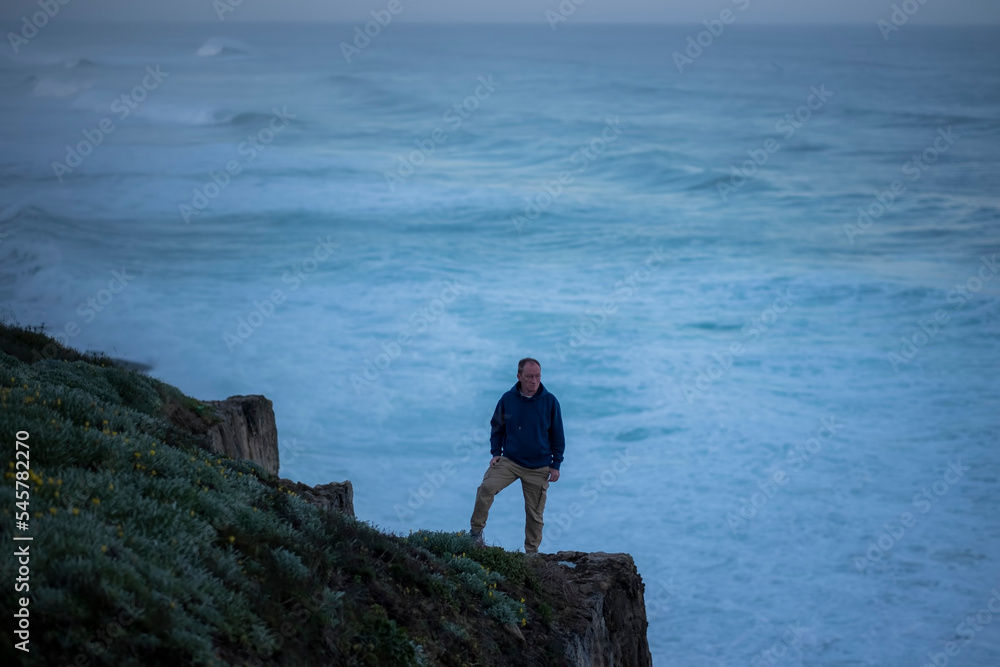 Obraz premium A man stands on the edge of a cliff against the morning ocean surf.