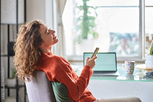 Young smiling pretty woman holding smartphone using cell mobile phone taking break relaxing while remote working or learning from home sitting on chair at table with laptop. photo