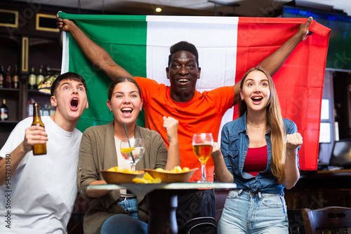 Multiracial Italy sports team fans, men and women, supporting their favourite team in bar, raising state flag and screaming chants together.