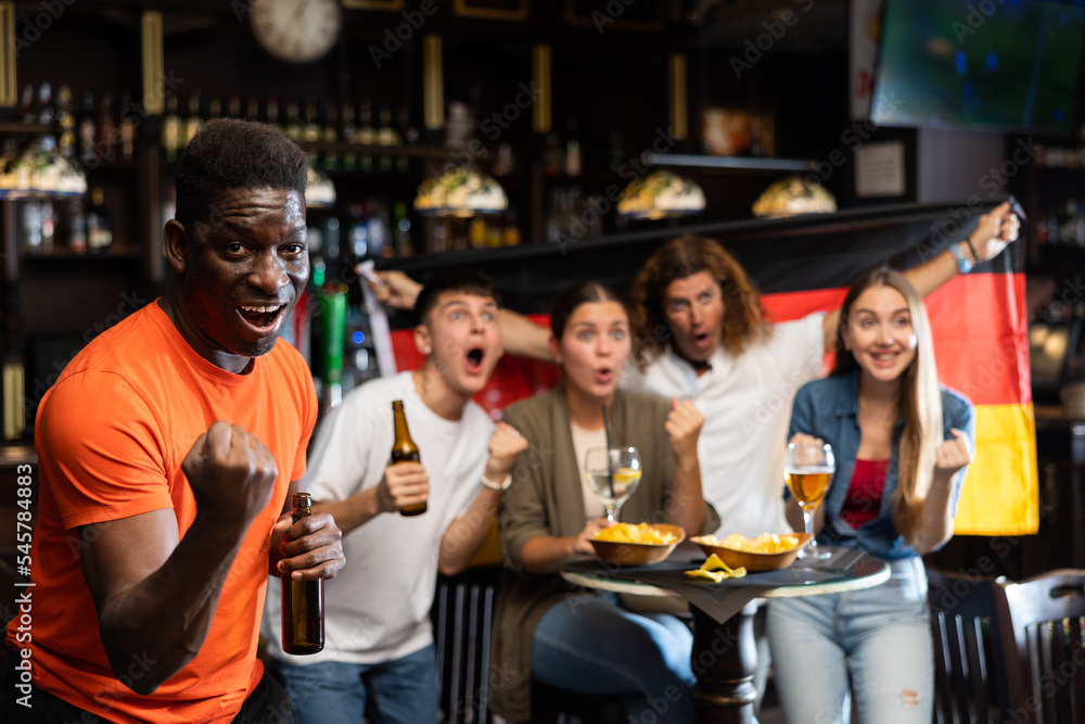 Happy excited african american man celebrating victory of favorite team while watching sports game in pub on background of friends holding flag of Germany