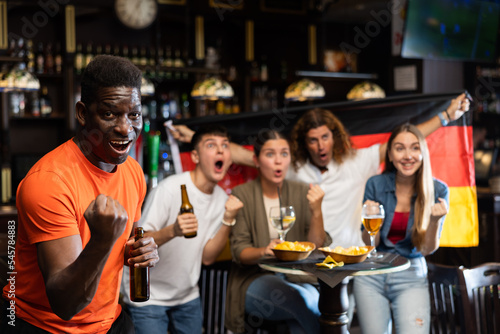 Happy excited african american man celebrating victory of favorite team while watching sports game in pub on background of friends holding flag of Germany