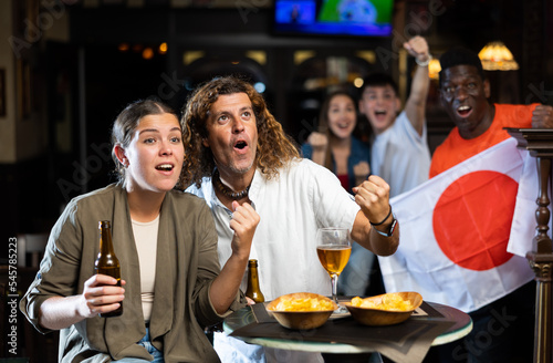 Happy diverse soccer fans with flag of Japan, rejoicing winning game with glasses of beer and snacks in the pub