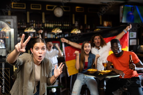 Cheerful excited young female fan watching sports game in bar, gesturing emotionally, happy with victory of favorite team against background of people with national flag of Germany..