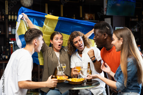 Excited multiracial footbal fans waving the flag of Sweden while drinking beer and watching tournament in the pub