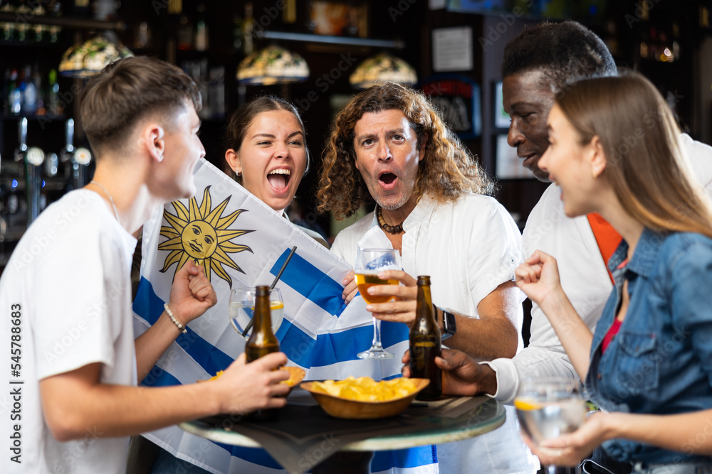 Group of Uruguay football team fans spending time in bar, drinking bear and having fun. People with state flag in pub. - obrazy, fototapety, plakaty 