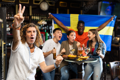 Male Sweden football team fan spending time in bar, gesturing and screaming chants. People with state flag in pub.