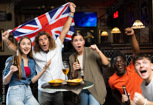 Excited young adults of different nationalities, sports fans celebrating victory of favorite team with beer in bar, waving national flag of United Kingdom..