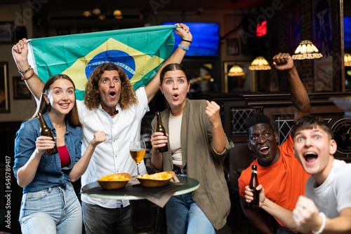 Emotional cheerful young adults friends, football fans cheering for favorite Brazil team together while watching match on tv in sports bar