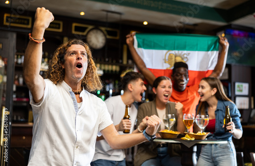Male Iran football team fan spending time in bar, making gestures and screaming chants. People with state flag in pub.