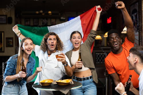 Group of young adult people, football fans of Italian team watching match on TV and emotionally rooting for victory in sports bar, waving national flag of Italy