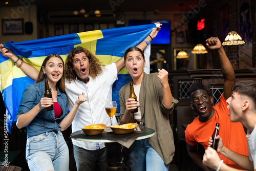 Group of happy emotional football fans having fun in sports bar, celebrating victory of favorite Swedish team together after watching match