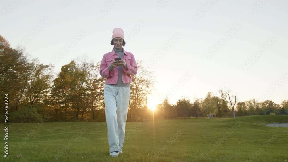 Beautiful, sunny day in colorful autumn park. Young, brunette woman in colorful outfit texting on her smartphone while strolling along lush, green meadow. High quality 4k footage Beautiful, sunny day in colorful autumn park. Young, brunette woman in colorful outfit texting on her smartphone while strolling along lush, green meadow. High quality 4k footage
