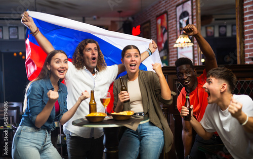 Excited diverse soccer supporters with flag of Russia watching tournament with pint of beer and chips in the pub