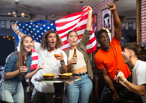 Group of emotional young adults, American football fans cheering for favorite team together in sports bar, waving flag of United States while watching match on TV