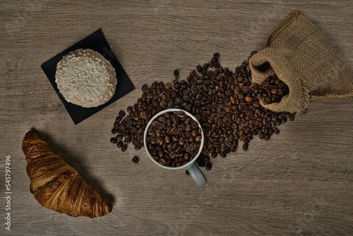 Top view of brown roasted coffee beans spilled on a table with a full coffee mug and biscuit