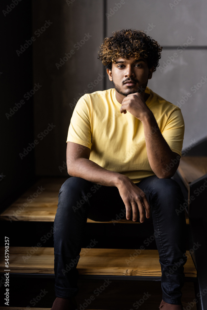 Thoughtful indian eastern ethnic man user sitting at home on stairs relaxing wearing earbuds using smartphone thinking looking through window drinking coffee listening music, audio book or podcast. Thoughtful indian eastern ethnic man user sitting at home on stairs relaxing wearing earbuds using smartphone thinking looking through window drinking coffee listening music, audio book or podcast.