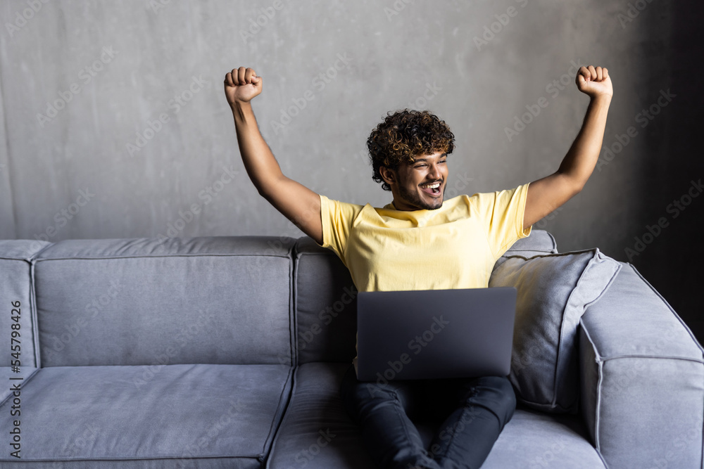 Successful young indian man lying on the couch while raising hands and using a laptop in the living room