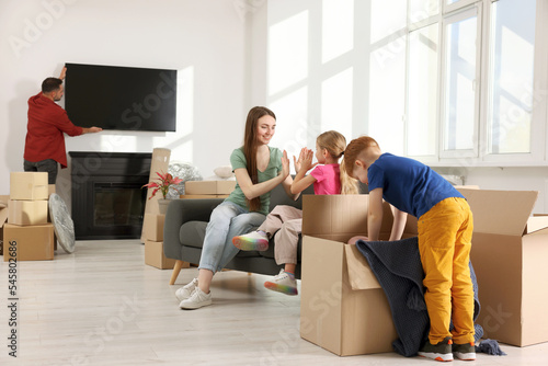 Family settling into new home. Dad setting up TV, happy mom and daughter playing on couch, son unpacking his boxes. Moving day