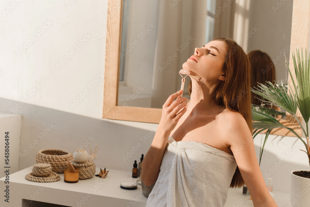 Young caucasian woman is massaging her neck with a massage roller to tighten, cool and soothe skin. Home spa concept. Young caucasian woman is massaging her neck with a massage roller to tighten, cool and soothe skin. Home spa concept.